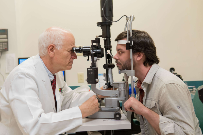 dr baker looks into a patient's eyes during an exam. the patient sits across from dr baker with his chin on a plastic stand