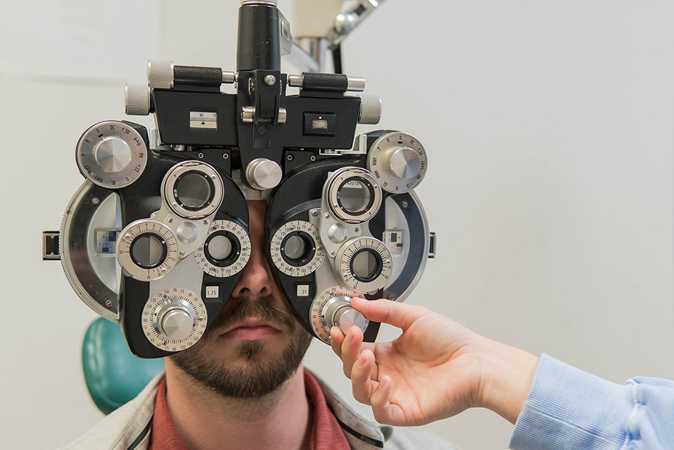 a hand adjusts a dial on an eye exam instrument as a patient sits behind it with his eyes looking through the glass