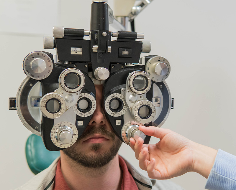 a hand adjusts a dial on an eye exam instrument as a patient sits behind it with his eyes looking through the glass