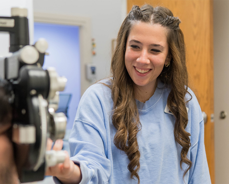 A woman with long hair smiles and adjusts an eye exam machine