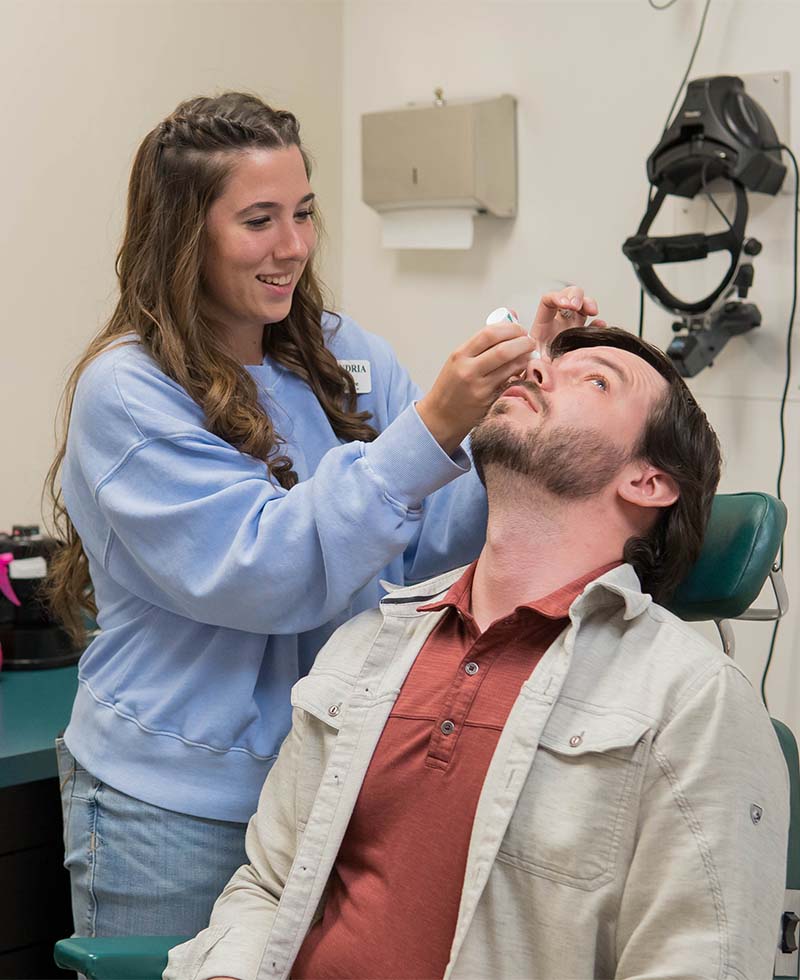 a nurse in a blue sweatshirt puts eyedrops into a patient's eye. the patient has his head back so the drops don't drop down his face.