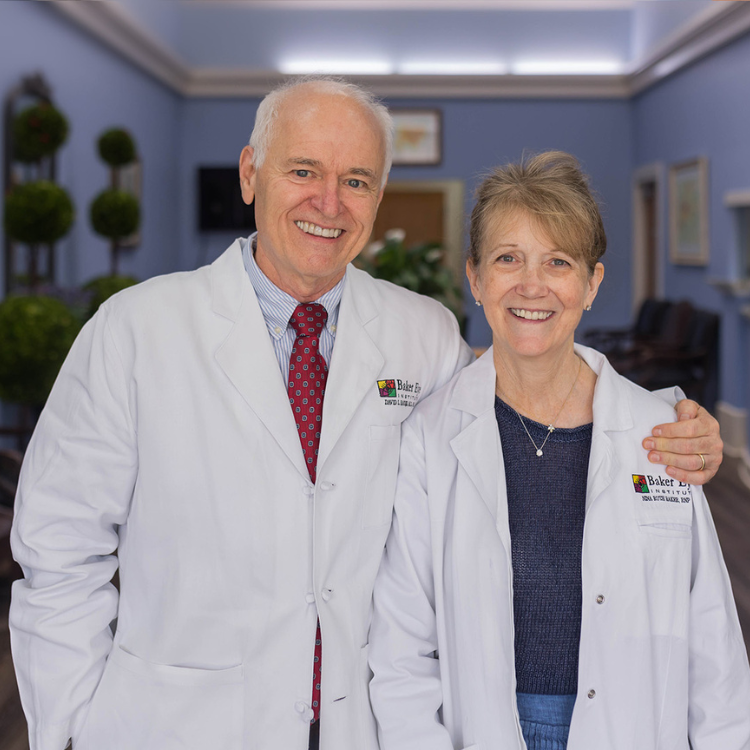 Dr Baker and Nina Baker, an older white couple, stand together each in white doctor coats, smiling in the blue waiting room of Baker Eye Care. Dr baker has his left arm around Nina.